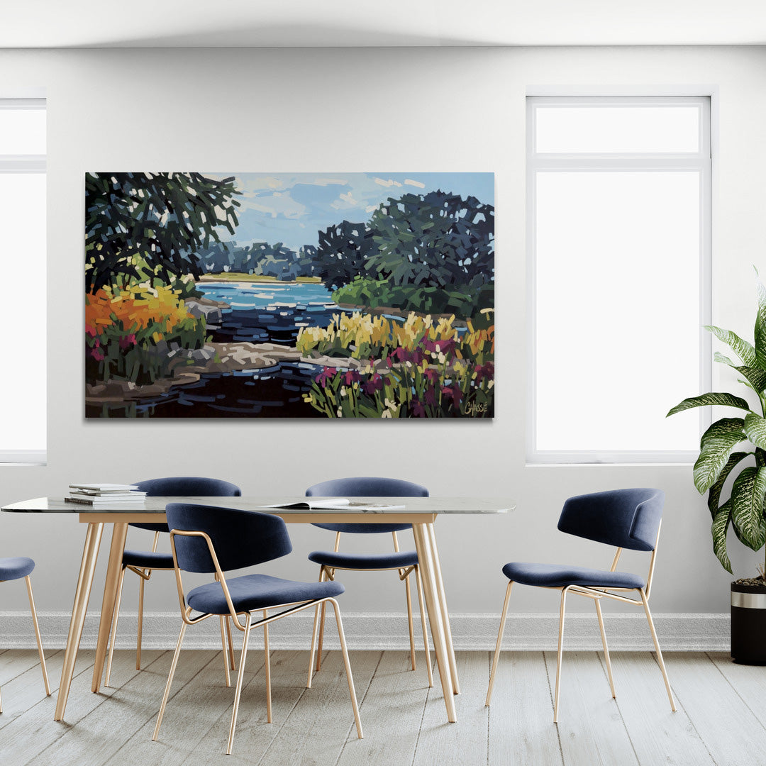 Une salle à manger lumineuse et moderne avec un grand tableau coloré accroché au mur blanc central. Le tableau représente un paysage de nature morte de style impressionniste avec une rivière, des arbres sombres et des fleurs sauvages jaunes et violettes au premier plan. Sous le tableau se trouve une table à manger en verre transparent entourée de chaises en velours bleu marine aux pieds dorés fins. La pièce possède un sol en parquet de bois clair et deux grandes fenêtres laissent entrer la lumière du jour d