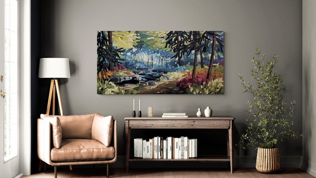 Toile de Martine Chassé accrochée au mur d’un salon moderne, au-dessus d’un meuble en bois, avec fauteuil, lampe et plante, représentant un paysage forestier lumineux traversé par un ruisseau.