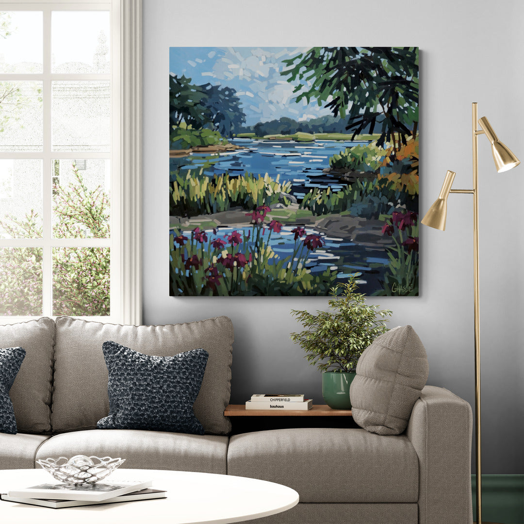 Grande peinture d'un paysage riverain calme accrochée au-dessus d'un canapé beige dans un salon lumineux de style scandinave contemporain. La scène naturelle aux reflets bleus et aux fleurs pourpres apporte une atmosphère paisible et contemplative à l'espace.